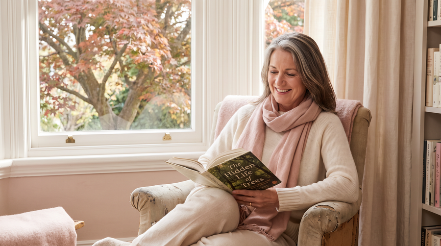 Smiling woman in her fifties in a cream sweater and blush scarf reading in a sunlit nook