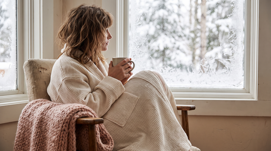 Woman in a cream waffle robe holding tea beside a frosted window in soft winter light