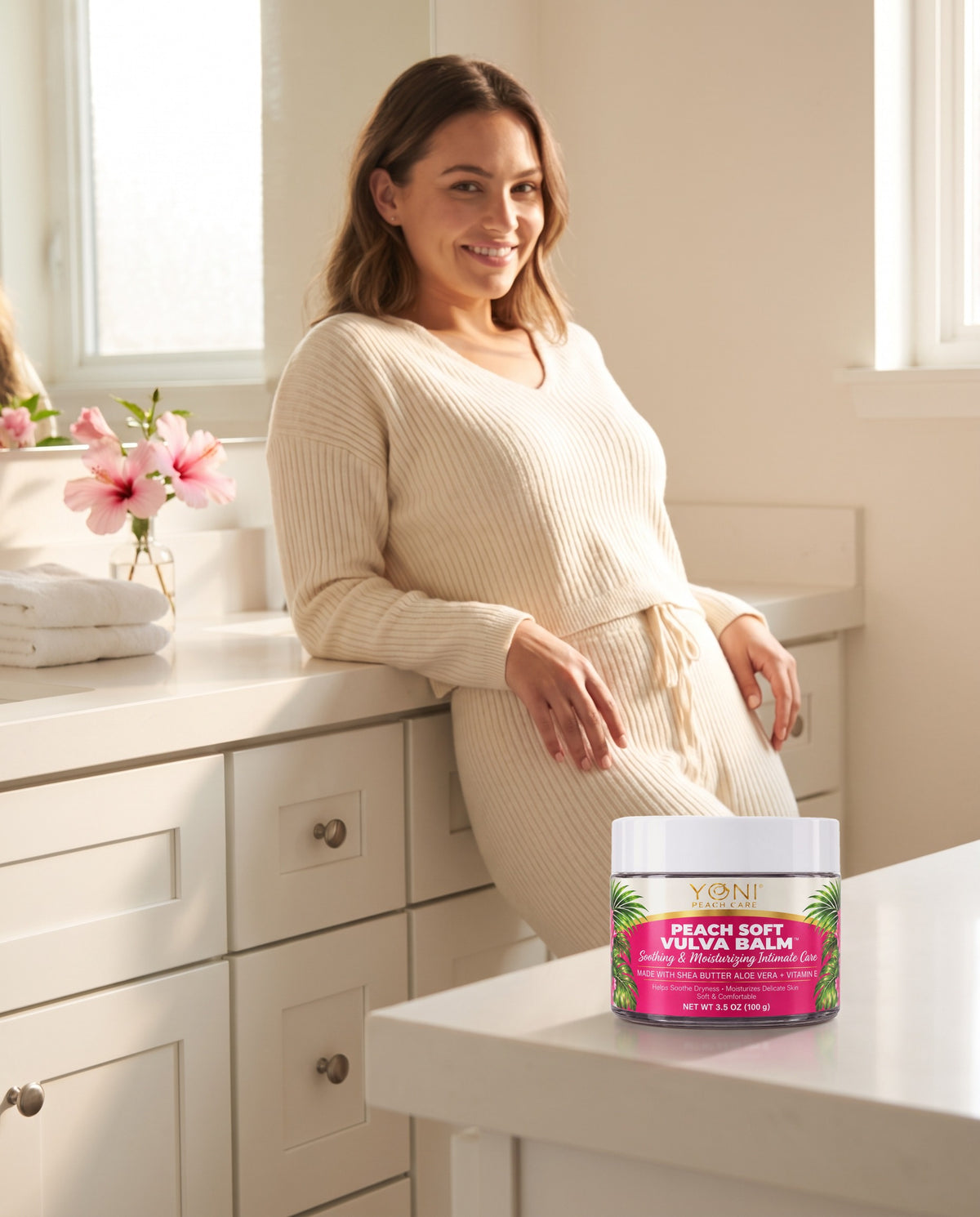 Woman in a cream knit lounge set at a bright bathroom vanity with Peach Soft Vulva Balm jar on the counter