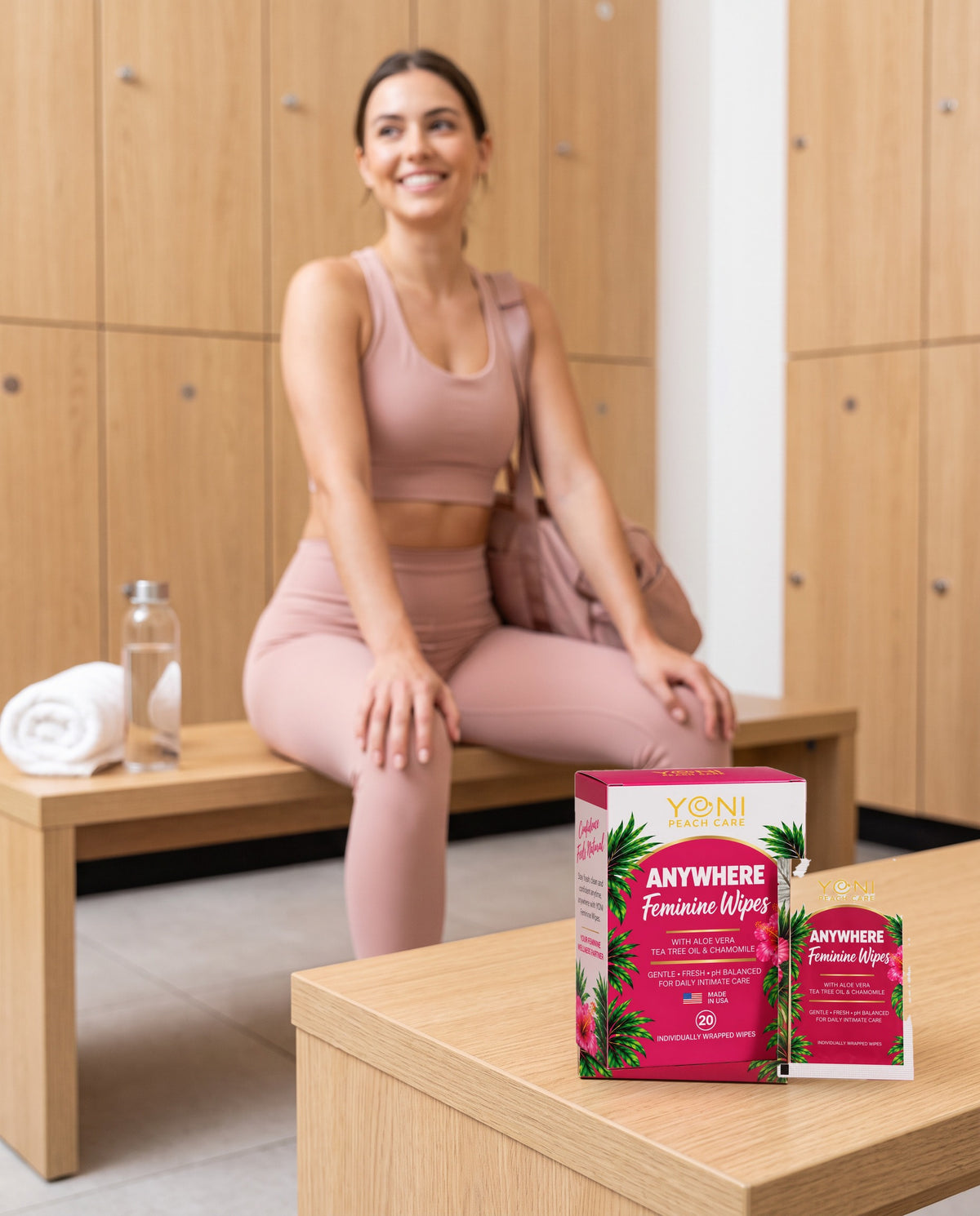 Woman in blush athleisure in a gym locker room with Anywhere Feminine Wipes box and sachet on the bench